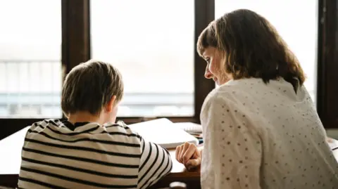 Getty/Maskot A boy and a woman sit in front of a table - their backs and the side of the woman's face are visible. The boy has brown hair and wears a black and white striped t-shirt, while the woman wears a white blouse. There are notebooks at the table and windows on the back wall.