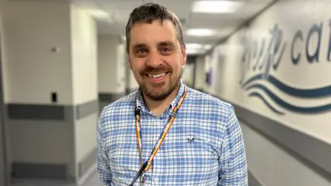 Jon Staple is standing in a hallway. He is wearing a blue and white shirt and wearing his NHS lanyard. He is smiling at the camera.