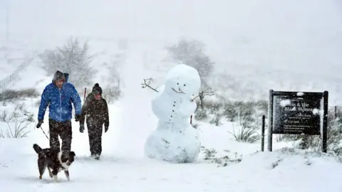 Getty Images A father and son walking their black and white dog on Black Mountain in the snow of December 2017. There is a large snowman next to them.