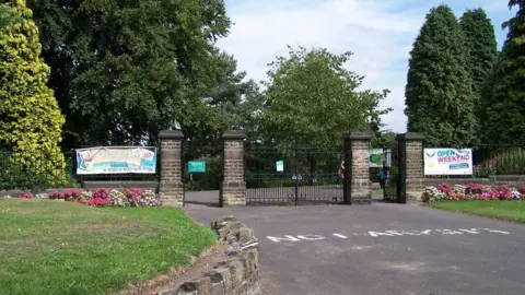 Geograph/Terry Robinson The gated entrance to Graves Park. There are four large stone pillars around 8ft tall with large cast iron gates. In front there is a tarmac road with raised verges on either side and borders of pink and white flowers.