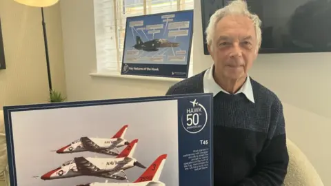 A man with white hair is sat on a cream chair, wearing a blue jumper with a white collar shirt underneath. He is holding a picture of red and white Hawk jets.