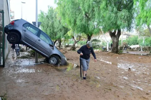 A woman using an umbrella for support walks next to a car lifted up on its front two wheels and balanced against a building in a street covered in mud in a flooded area in Picanya, near Valencia.