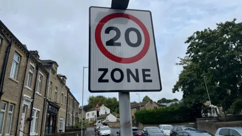 A road sign for a 20mph speed zone in a residential area, with houses to the left and cars on the road.