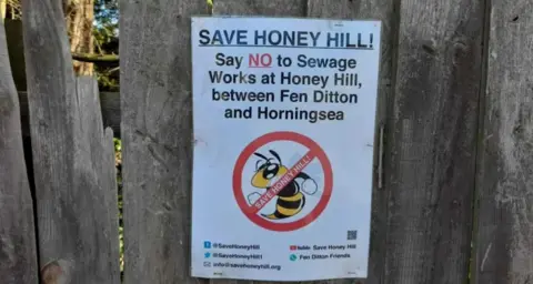 A poster on the wooden fence. The poster reads "Save Honey Hill! Say NO to sewage works at Honey Hill between Fen Ditton and Horningsea". The text is accompanied by an image of a yellow and black bee, with a red circle and line through the image.  