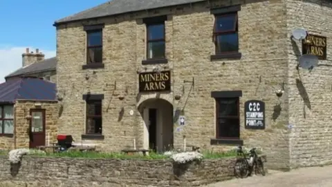 The exterior of the Miners Arms, a brick building with prominent signage and a flower bed to the front. A bike is parked up against a low wall, under a sign saying 'C2C Stamping Point'.