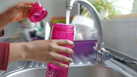 A woman fills up a pink bottle with water from a kitchen tap in a bright kitchen.