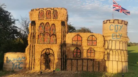 A sculpture of Ely Cathedral made out of straw bales against a field backdrop, with a Union Flag flying from the Octagon Tower