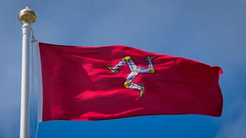 MANX SCENES An Isle of Man Flag flying on a flagpole against a blue sky. The flag is red, with a symbol of three armoured legs in the centre. 