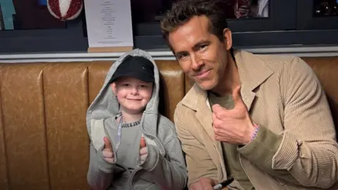 Riley, a young boy wearing a cap and a black hoodie, and Ryan Reynolds, who wears a cord jacket. They are both giving thumbs up to the camera.