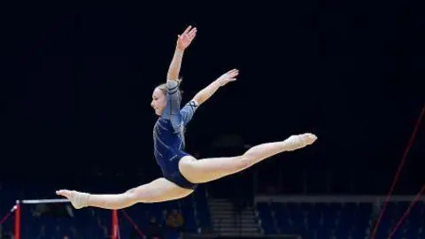 Justyna Kosinink Olivia, performs a split leap on the floor exercise mat, with her arms extended gracefully. Gymnastics equipment, including uneven bars, is visible in the background, along with spectators and judges in a large arena setting.