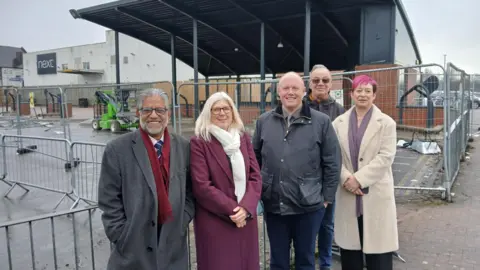 Five people stand in front of railings around a shelter at a bus station. There are three men and two women in the group, all of whom smile at the camera. The silver railings surround a tall black overhead canopy, and a white building with a black Next sign is behind the shelter.