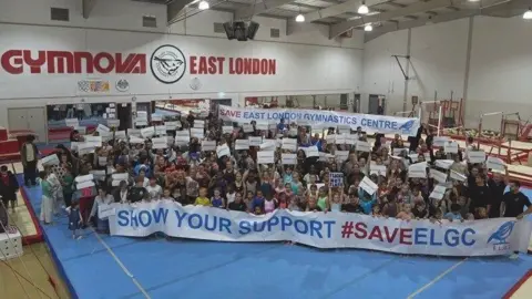 ELGC A large group of people, including children and adults, gather in a gym holding signs in support of saving the East London Gymnastics Centre. They stand behind a banner reading, "SHOW YOUR SUPPORT #SAVEELGC." The gym has "East London" and "Gymnova" signage in the background.