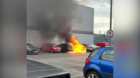 A fire spreading to parked cars. The blaze is burning bright, with the fire engulfing a dark vehicle to the left of the main fire. 