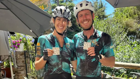 A man and a woman wearing white bike helmets and matching blue and black cycling gear smile and hold up two white medals. There's lush green vegetation and blue sky behind them.