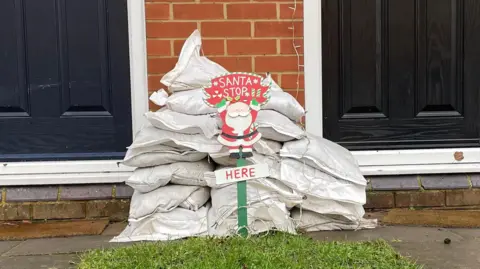 Ben Schofield/BBC A 'Santa stop here' sign in front of a pile of sandbags. There is a small patch of grass visible in front of the sign. Behind the sandbags the bottom half of two front doors can be seen.