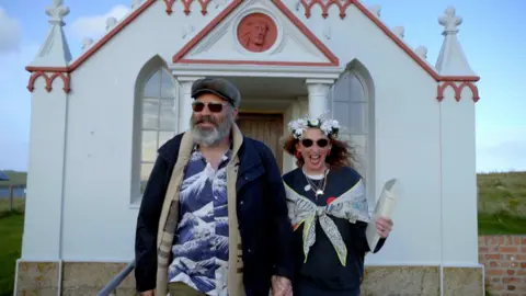 Greg has a grey beard and wears sunglasses and dark blue shirt with wave patterns, while Jules has curly blonde hair and wears a flower crown and shawl. They are walking out of white chapel with terracotta cornicing hand in hand. Jules holds their marriage renewal certificate in her left hand. Both are smiling.