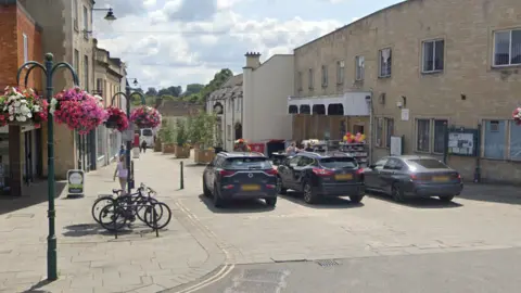 A Google street view of Calne High Street, showing cars parked at one end and planters with trees running down the street