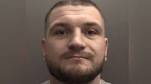 A police custody photograph of Martin Collins. He has short cropped and shaven dark-brown hair with same coloured beard and brown eyes. He is staring into the camera.