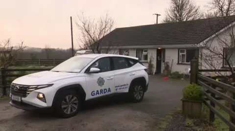 A Garda car sits in front of a white bungalow. A Christmas decoration hangs from the front door. A pair of little trees sit on each side of the front door. 