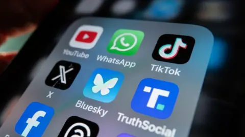 An iPhone screen is lit up and shows an open folder with various social media logo apps including YouTube, WhatsApp, TikTok, X, Bluesky, TruthSocial and Facebook