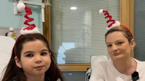 Lis, wearing festive pyjamas with gingerbread men and snowmen, sits beside her mum Rebecca, both wearing red spiral Santa hat headbands, in a hospital room at Great Ormond Street Hospital.