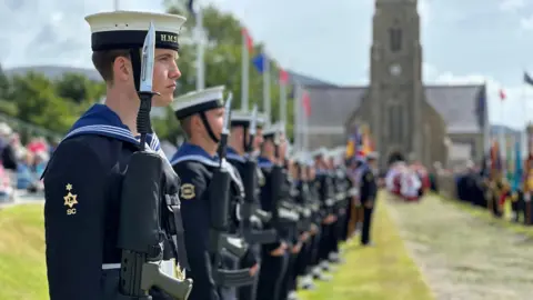 A young man in a Royal Navy Uniform with a gun on his shoulder heads the row of the Guard of Honour, which is lining the ceremonial walkway. Members of the clergy dressed in white and red robes can be seen in the background walking up the walkway with Manx flags flying on either side and the Royal Chapel in the background.