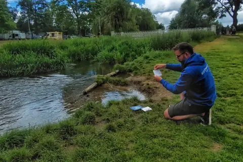 A man wearing a blue hoodie with the slogan Earthwatch kneeling on a riverbank holding up a science card that shows the chemical levels in the river. The river is small and flowing through clumps of grasses