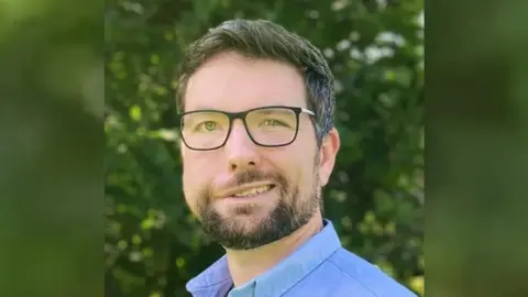 Aaron Jones with dark hair and glasses smiling at the camera. He is standing in front of a bushes