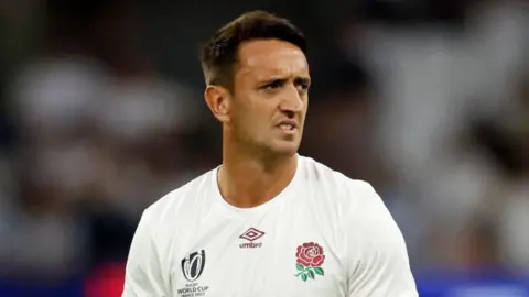 Alex Mitchell in a white England Rugby training top. He is looking to the side a pulling a quizzical face.