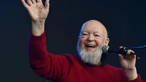 Reuters Sir Michael Eavis in a red jumper holding a microphone on stage.