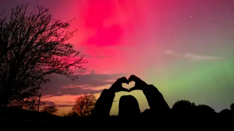 A silhouette of a person holding up a love heart directly towards a backdrop of the Northern Lights in bright red, bright green and darker orange and purple tones