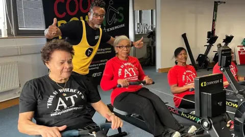 Five Cool Rowings members wearing Ageless Teenagers T-shirts on rowing machines. Weininger stands in the back with his thumbs up. 