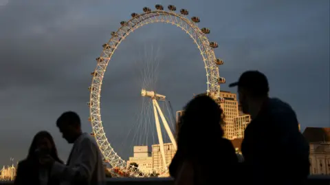 London Eye illuminated by sunlight in background under cloudy skies with silhouettes of people in foreground