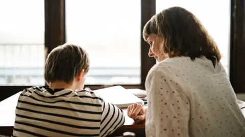 The back of a child wearing a stripey T-shirt sat at a table with books in front of him, with a woman sat next to him wearing a white blouse, turning slightly so a little of her side profile can be seen.