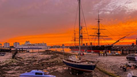 MONDAY - A ferry passes behind HMS Warrior under this vivid Portsmouth sunset. The sky is glowing a deep orange that is reflected in the mud of Portsmouth Harbour. The ferry passing is white and behind you can see the tower blocks in Gosport. In the foreground is a small sailing boat that is in front of HMS Warrior with its black and red hull and masts.