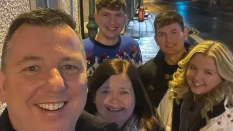 Glenn Johnson Glenn Johnson's selfie with his wife and children after they all helped to serve last year's Christmas Day dinner.  Glenn and his two sons all have short, dark hair and they are wearing Christmas jumpers. His wife and daughter have shoulder-length hair and they are wearing pale-coloured coats. 
 