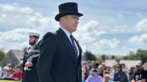 Alfred Cannan wearing a dark suit and top hat walking part the gathered crowds towards Tynwald Hill.