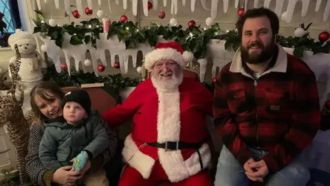 Three-year-old Ralf admires the Christmas decorations as he meets Santa for the first time without shielding. Father Christmas in sat in the middle, with one arm around Daisy, who is cuddling Ralf, who is looking up and dressed in a black beanie and green winter coat and gloves, and another arm around Ralf's dad Doug, dressed in a red and black checked shirt and jeans. Strings of baubles and fake icicles are behind them