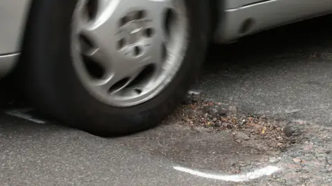 PA Media A silver car driving over a pothole on the road that has white spray paint markings around it.