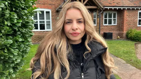 A woman with long blonde hair and wearing a black coat and black top, stands in front of a brick house. She is in the front garden with a concrete path to one side and lawns either side. A large green bush is to her right.