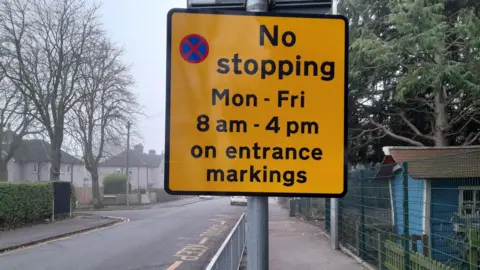 LDRS A yellow sign showing parking restrictions outside a school, with keep clear road markings on the road