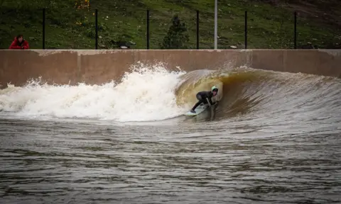 A young surfer with a green helmet rides through a crashing white wave. A person in a red jacket watches over a brown wall.