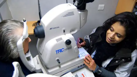 Getty Images A patient looks into an eye scanner, while a health work looks on.