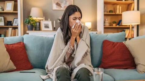 A woman sitting on her couch sneezing into a handkerchief. She has a shawl wrapped around her. She has dark hair, while a glass of water sits on a table in front of her. Bookcases and cabinets are visible behind her. 