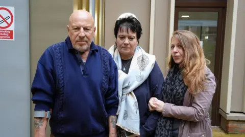 Shaun Whitmore/BBC Holly Goodchild's family posing for a picture outside a court room at Norfolk Coroners' Court, including her dad - a bald man with tattoos and a crutch wearing a navy jumper - his partner, a woman with short black hair wearing a large white scarf over a navy top, and Holly's sister - a young woman with shoulder-length blonde hair, wearing a light purple jacket and a black and grey scarf.