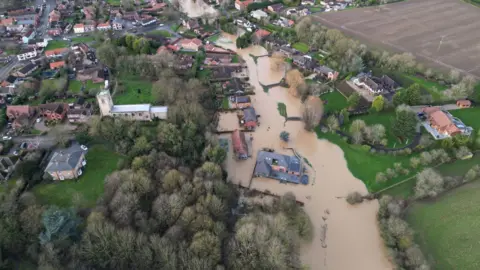 Laura Calvert An aerial view of the street, which is partly under water. On the left is housing and a church, surrounded by green land and trees, while on the right is more housing and a stretch of farmland 