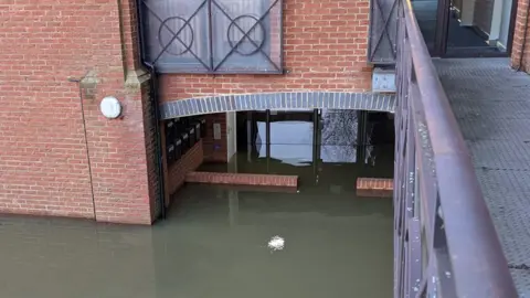 Richard Ayres A shot looking down on to flood waters surrounding a red brick, relatively modern-looking building. The dark green water is several bricks high and can be seen reaching underneath an arched area, through some doors and into a lobby, which appears to also be underwater. A row of mail boxes are just visible underneath the arch and are around six bricks above the water. A raised metal pathway and handrail can be seen on the right of the image. 