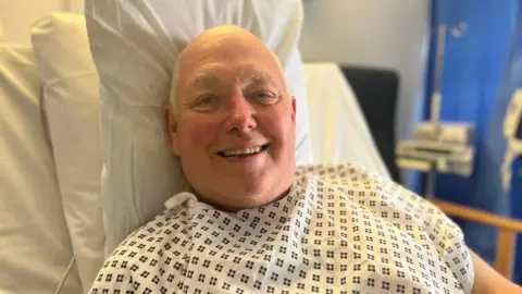 David Ince propped up on a hospital bed and smiling towards the camera. He is wearing a white and black hospital robe. He has short white hair and is smiling. 