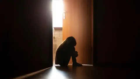 Getty Images Despairing child in silhouette sitting with head on knees in the dark frame of a doorway, backlit by a room behind flooded with daylight - stock photo.