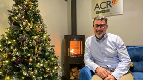 Jason Searle sits next to one of their wood-burning stoves. To the left is a Christmas tree and above his head is the company's sign. He has grey, white hair and a grey, white beard and wears glasses. He has a blue shirt on with light brown trousers and sites on a blue sofa. 